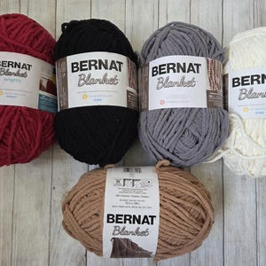 May include: Five skeins of Bernat Blanket yarn in various colors: red, black, gray, cream, and tan. Each skein has a label with the brand name and product details. The yarn is thick and soft, ideal for blankets and other cozy projects.