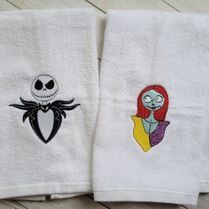 Pode incluir: Duas toalhas de felpa brancas com desenhos bordados. A toalha da esquerda mostra uma personagem em preto e branco com rosto esquel&eacute;tico e fato. A toalha da direita apresenta uma personagem com cabelo ruivo, pele azul clara e vestido de retalhos.