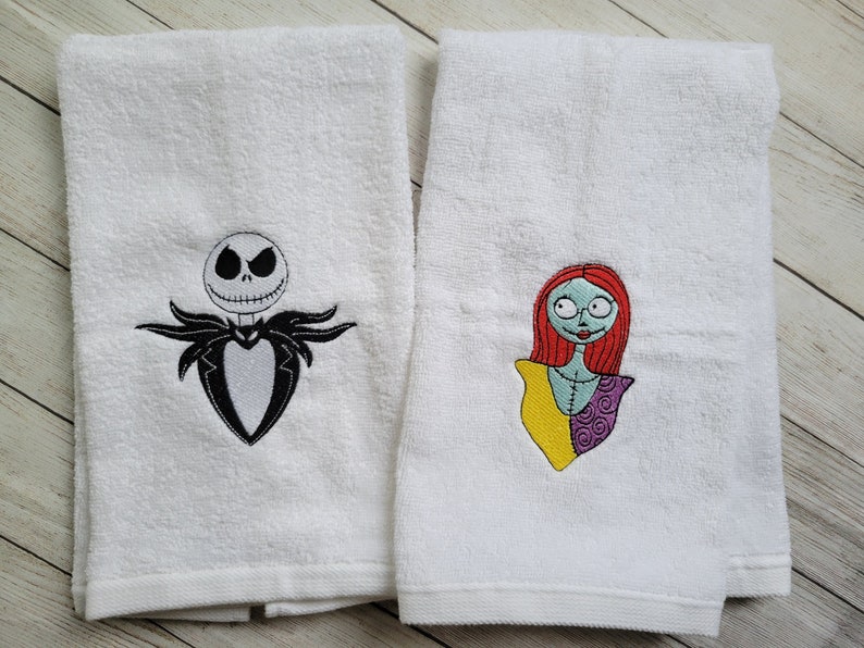 Pode incluir: Duas toalhas de felpa brancas com desenhos bordados. A toalha da esquerda mostra uma personagem em preto e branco com rosto esquel&eacute;tico e fato. A toalha da direita apresenta uma personagem com cabelo ruivo, pele azul clara e vestido de retalhos.