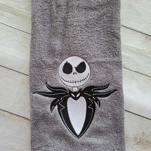 Pode incluir: Uma toalha de m&atilde;o cinzenta com um design bordado de uma personagem esquel&eacute;tica com cabe&ccedil;a redonda branca, olhos pretos vazios e boca costurada. A personagem veste um casaco preto com frente de camisa branca e la&ccedil;o em forma de morcego.
