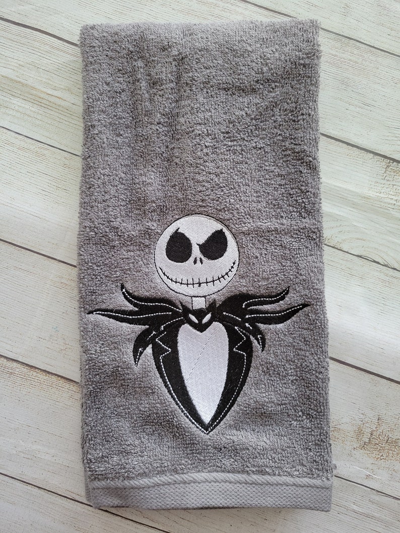 Pode incluir: Uma toalha de m&atilde;o cinzenta com um design bordado de uma personagem esquel&eacute;tica com cabe&ccedil;a redonda branca, olhos pretos vazios e boca costurada. A personagem veste um casaco preto com frente de camisa branca e la&ccedil;o em forma de morcego.