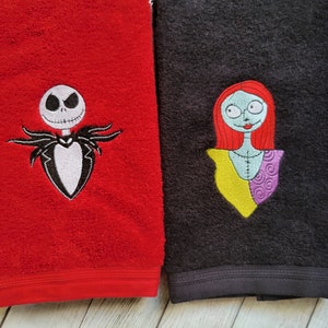 Pode incluir: Uma toalha vermelha com um bordado da personagem Jack Skellington e uma toalha preta com um bordado da personagem Sally. Ambas as toalhas t&ecirc;m uma superf&iacute;cie de tecido texturizado e um detalhe de faixa horizontal perto da borda inferior.