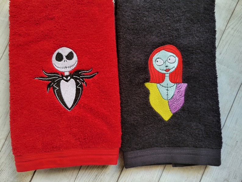 Pode incluir: Uma toalha vermelha com um bordado da personagem Jack Skellington e uma toalha preta com um bordado da personagem Sally. Ambas as toalhas t&ecirc;m uma superf&iacute;cie de tecido texturizado e um detalhe de faixa horizontal perto da borda inferior.