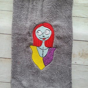 Pode incluir: Uma toalha de m&atilde;o cinzenta com um bordado de uma personagem com cabelo comprido vermelho, pele azul clara e detalhes costurados. A personagem veste uma pe&ccedil;a amarela e roxa com padr&otilde;es. O bordado est&aacute; centrado no tecido texturizado.