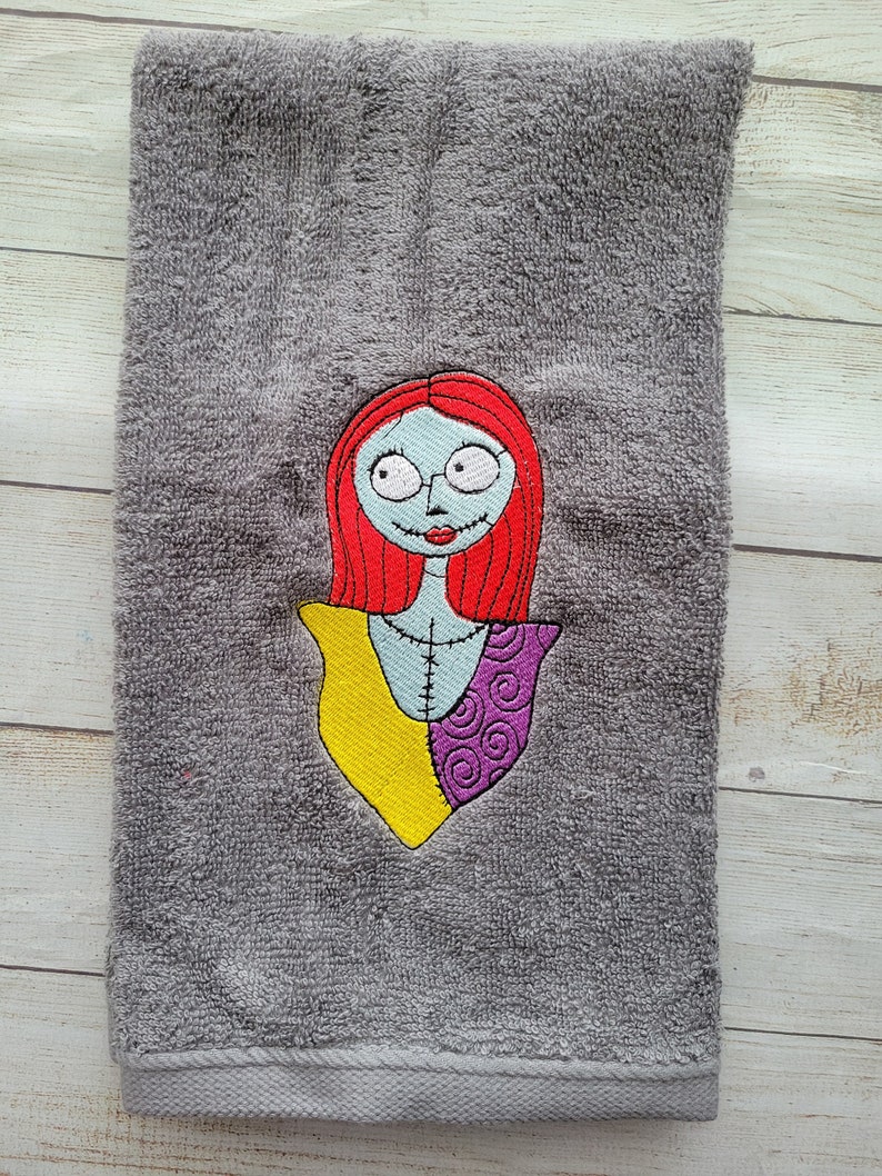 Pode incluir: Uma toalha de m&atilde;o cinzenta com um bordado de uma personagem com cabelo comprido vermelho, pele azul clara e detalhes costurados. A personagem veste uma pe&ccedil;a amarela e roxa com padr&otilde;es. O bordado est&aacute; centrado no tecido texturizado.