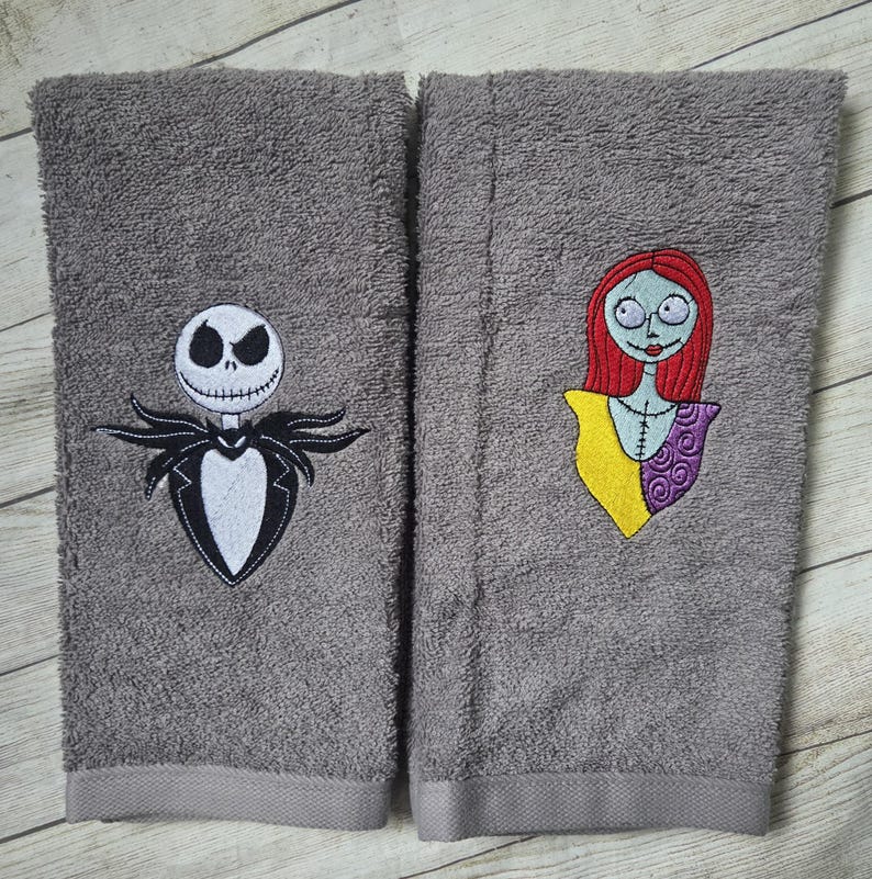 Pode incluir: Duas toalhas de m&atilde;o retangulares cinzentas com desenhos bordados. A toalha da esquerda mostra uma figura branca com cara de caveira num fato preto. A da direita apresenta uma personagem com cabelo vermelho, top amarelo e manga roxa.