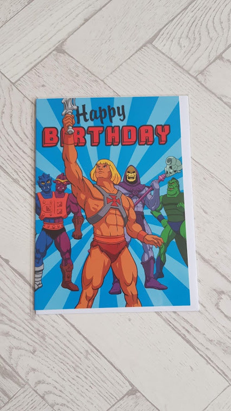 Puede incluir: Una tarjeta de cumplea&ntilde;os con He-Man y otros personajes de la serie animada Masters of the Universe. La tarjeta tiene un fondo azul con un mensaje "Feliz cumplea&ntilde;os" en rojo y blanco.
