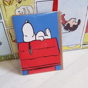 K&ouml;nnte beinhalten: Eine Gru&szlig;karte mit einem Comic-Hund, Snoopy, der auf seinem roten Hundehaus liegt. Die Karte hat einen blauen Hintergrund und der Text "Charlie Brown" ist im Hintergrund sichtbar.