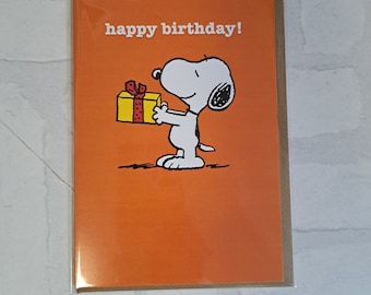 Può includere: Un biglietto d'auguri arancione con un'illustrazione in bianco e nero di Snoopy che tiene in mano un regalo giallo con un nastro rosso. Il testo "happy birthday!" è stampato in lettere bianche in alto sul biglietto, inserito in una busta di plastica trasparente.