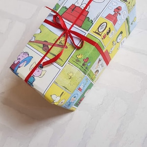 K&ouml;nnte beinhalten: Ein Geschenk, das in wei&szlig;es Papier mit einem roten Band eingewickelt ist. Das Papier zeigt ein Comic-Design mit Figuren aus dem Peanuts-Comic. Der Text auf dem Papier lautet "Good Grief".