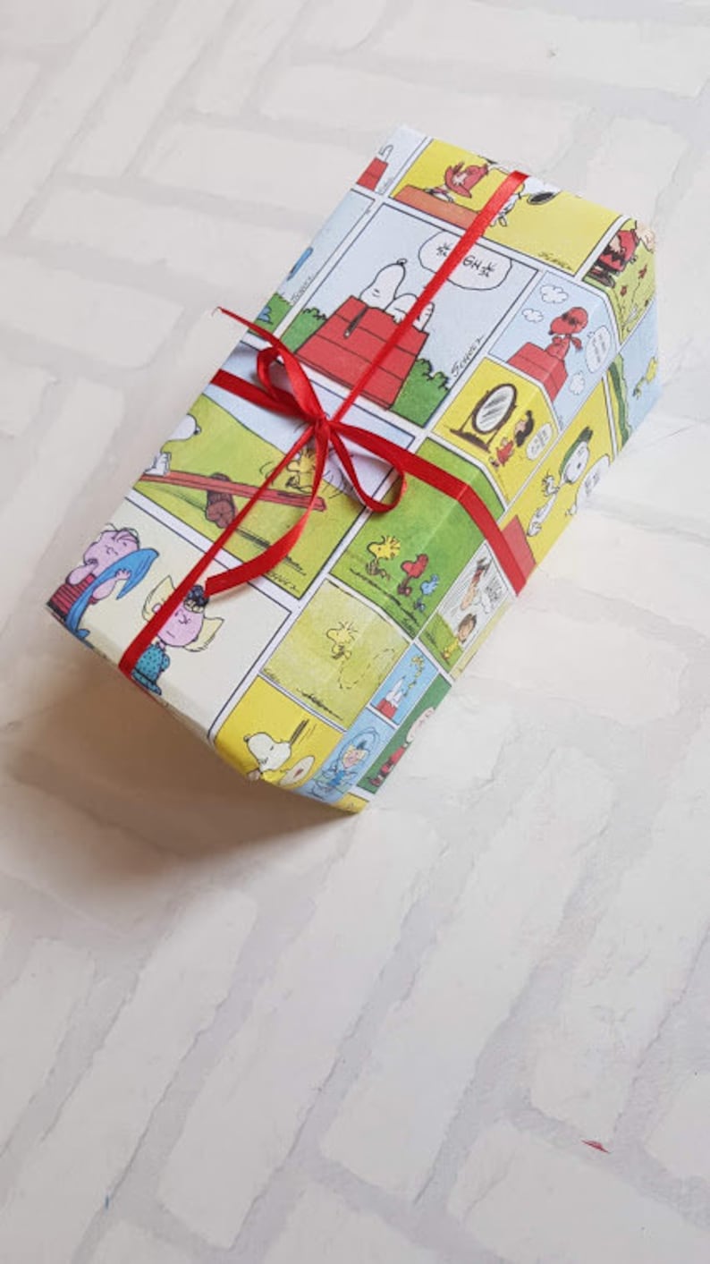 K&ouml;nnte beinhalten: Ein Geschenk, das in wei&szlig;es Papier mit einem roten Band eingewickelt ist. Das Papier zeigt ein Comic-Design mit Figuren aus dem Peanuts-Comic. Der Text auf dem Papier lautet "Good Grief".
