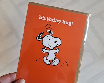 Può includere: Biglietto di auguri di compleanno arancione con un'immagine di Snoopy che fa un abbraccio. La carta dice "birthday hug!" in testo bianco.