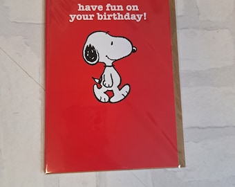 Può includere: Biglietto di auguri di compleanno rosso con un personaggio dei cartoni animati Snoopy bianco e il testo "have fun on your birthday!"