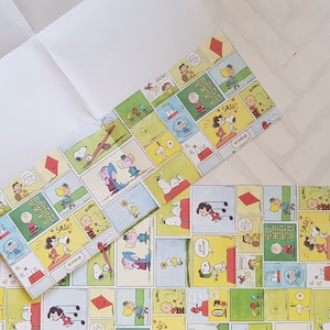 K&ouml;nnte beinhalten: Ein Geschenkpapier mit einem Peanuts-Comic-Design. Das Papier hat einen gelben Hintergrund mit roten, blauen und gr&uuml;nen Comic-Feldern. Die Felder zeigen Figuren wie Snoopy, Charlie Brown und Lucy. Der Text in den Feldern lautet "Hee! Hee! Hee!" und "Smak!"