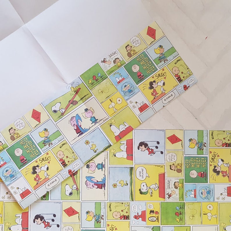 K&ouml;nnte beinhalten: Ein Geschenkpapier mit einem Peanuts-Comic-Design. Das Papier hat einen gelben Hintergrund mit roten, blauen und gr&uuml;nen Comic-Feldern. Die Felder zeigen Figuren wie Snoopy, Charlie Brown und Lucy. Der Text in den Feldern lautet "Hee! Hee! Hee!" und "Smak!"