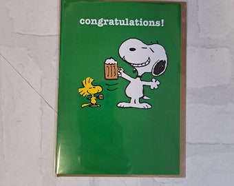 Può includere: Un biglietto di auguri verde con un'immagine di cartone animato di Snoopy e Woodstock. Snoopy tiene un boccale di birra e Woodstock è in piedi accanto a lui. Il biglietto dice "Congratulazioni!" in testo bianco.