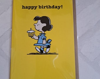 Può includere: Biglietto di compleanno giallo con un'illustrazione a cartoni animati di una ragazza che tiene in mano un cupcake con una candela accesa. Le parole "happy birthday!" sono stampate in nero in alto. Il biglietto è in una custodia di plastica trasparente.