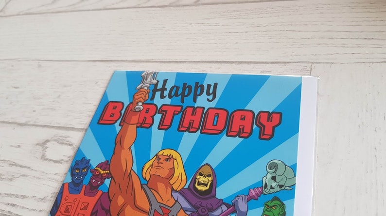 Puede incluir: Una tarjeta de cumplea&ntilde;os azul y roja con He-Man y los Masters del Universo. La tarjeta dice "Feliz cumplea&ntilde;os" en letras rojas.