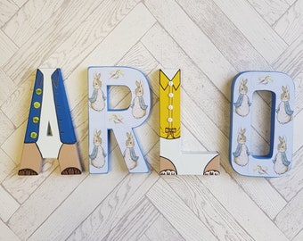 May include: Wooden letters spelling "ARLO" in a whimsical style. The "A" is blue with gold buttons, the "R" has a bunny pattern, the "L" is yellow with a bow tie, and the "O" has a bunny pattern. The letters are on a light gray background.