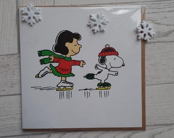 May include: A square greeting card featuring a cartoon illustration of Lucy and Snoopy ice skating. Lucy wears a red jumper and green scarf, while Snoopy wears a red hat. Three white glittery snowflake embellishments are attached to the card.