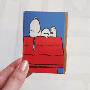 K&ouml;nnte beinhalten: Eine Gru&szlig;karte mit einer Schwarzwei&szlig;illustration von Snoopy, der auf seinem roten Hundehaus liegt, vor einem blauen Himmel.
