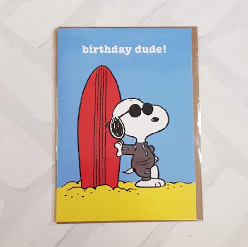 May include: Birthday card featuring Snoopy wearing sunglasses and a brown outfit, leaning against a red surfboard. The card has a blue background with the words "birthday dude!" printed in white. The bottom of the card shows yellow sand.