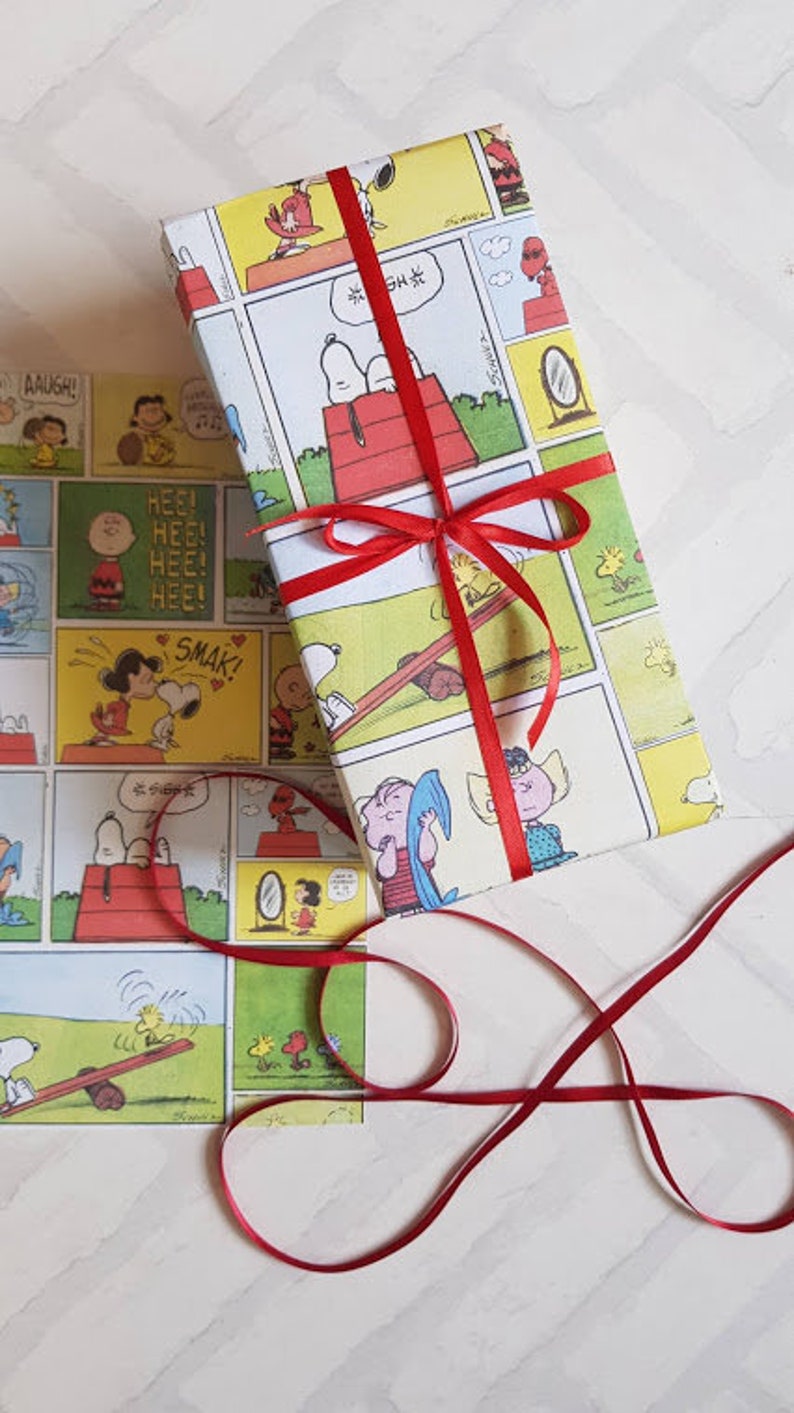 K&ouml;nnte beinhalten: Ein rotes Band, das um ein Geschenk gebunden ist, das in wei&szlig;es Papier mit einem Peanuts-Comic-Design eingewickelt ist. Der Comic zeigt Snoopy, Charlie Brown und andere Figuren. Das Papier ist gelb mit schwarzen Umrissen und roten Akzenten.