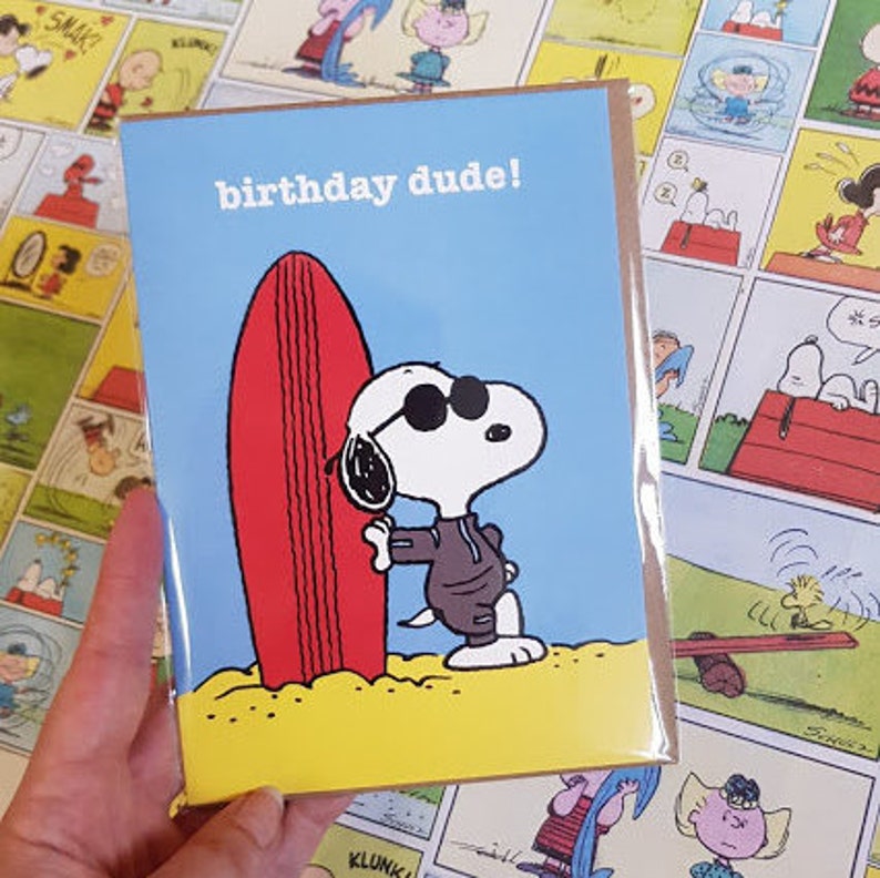 May include: Greeting card featuring Snoopy in sunglasses, holding a red surfboard on a beach. The card has the text "birthday dude!" against a blue sky background. The card is in a clear plastic sleeve.