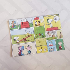 K&ouml;nnte beinhalten: Ein buntes Geschenkpapier mit einem Comic-Strip von Snoopy und der Peanuts-Gang. Das Papier zeigt verschiedene Szenen, darunter Snoopy beim Drachenfliegen, Snoopy auf einer Wippe und Snoopy beim Handstand. Das Papier ist gelb, gr&uuml;n, rot und blau.