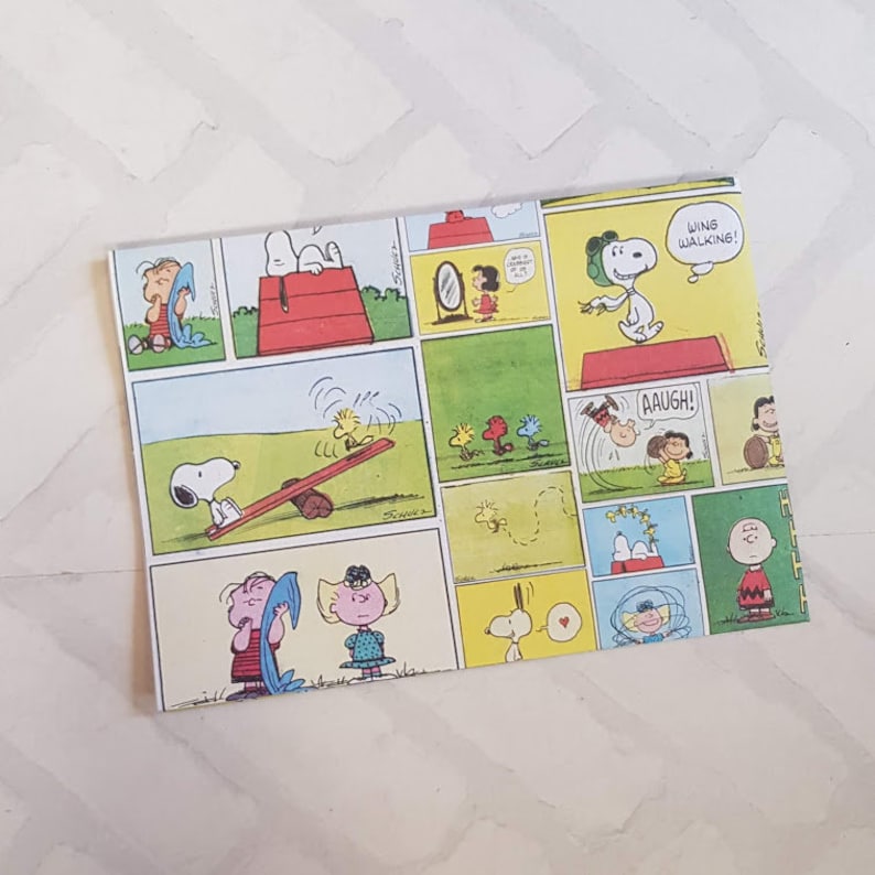 K&ouml;nnte beinhalten: Ein buntes Geschenkpapier mit einem Comic-Strip von Snoopy und der Peanuts-Gang. Das Papier zeigt verschiedene Szenen, darunter Snoopy beim Drachenfliegen, Snoopy auf einer Wippe und Snoopy beim Handstand. Das Papier ist gelb, gr&uuml;n, rot und blau.