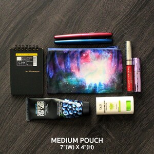 May include: A collection of beauty and stationery items on a dark wood surface. Includes a black notebook, two pens, a cosmetic pouch with a forest scene, lipsticks, a hand cream, and a sun stick. The pouch is 17.8 cm wide and 10.2 cm high.