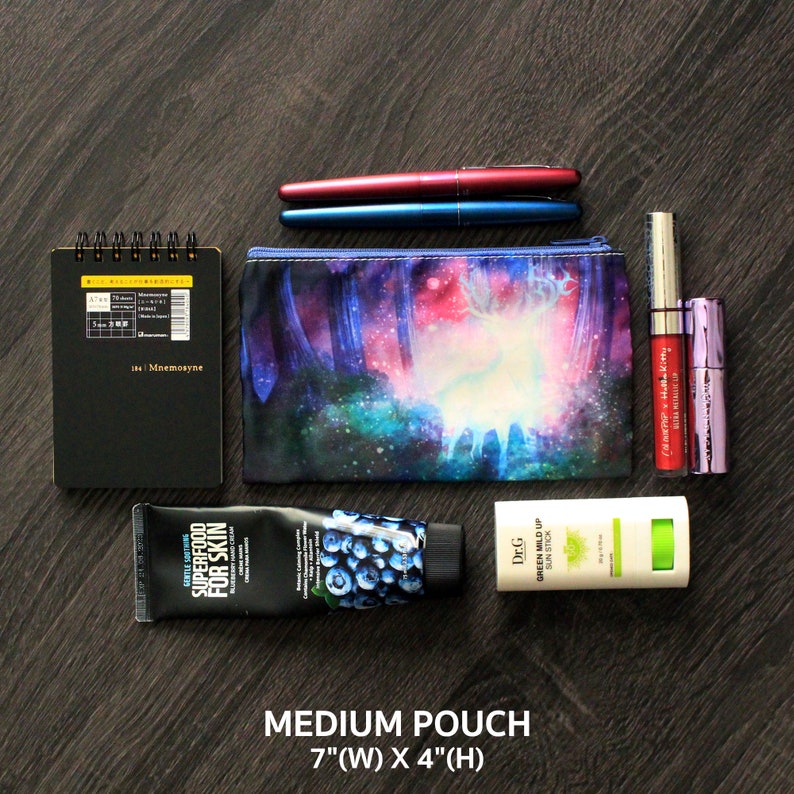May include: A collection of beauty and stationery items on a dark wood surface. Includes a black notebook, two pens, a cosmetic pouch with a forest scene, lipsticks, a hand cream, and a sun stick. The pouch is 17.8 cm wide and 10.2 cm high.