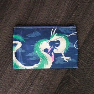 May include: A rectangular, navy blue zip pouch featuring a white and green dragon illustration against a cloudy sky background. The dragon is in a dynamic pose, with a person in silhouette. The zip is navy blue with white stitching.
