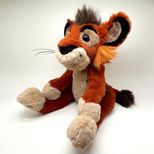 May include: A plush lion cub toy in shades of orange, brown, and tan. The toy has a detailed face with black accents, long whiskers, and a tuft of dark brown hair. The lion is sitting with its legs extended.