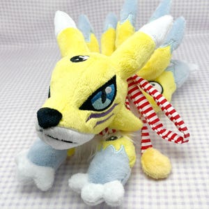 May include: Yellow and blue plush toy with nine tails, resembling a fox. The toy has a red and white striped scarf around its neck.