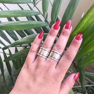 May include: Two silver rings with a wide band and a textured surface. The rings are worn on the index and middle fingers of a hand with red nail polish.