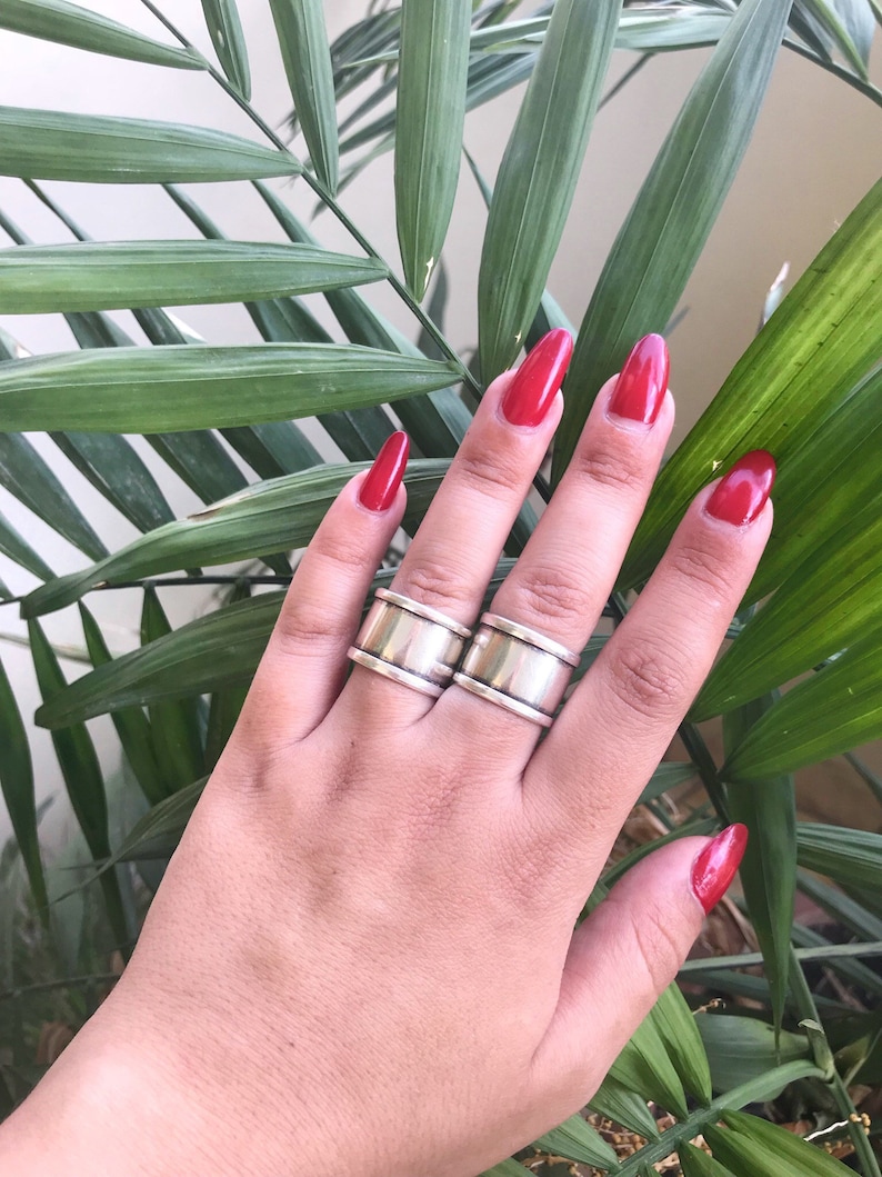 May include: Two silver rings with a wide band and a textured surface. The rings are worn on the index and middle fingers of a hand with red nail polish.