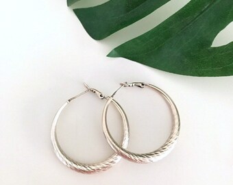 May include: A pair of silver hoop earrings with a textured finish. The earrings are large and have a simple, elegant design.