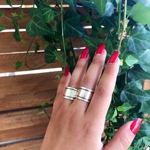 May include: Two silver rings with a wide, flat band. The rings are worn on the middle finger of a hand with red nail polish.