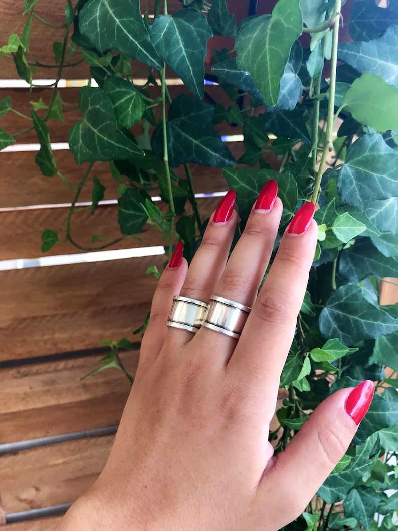 May include: Two silver rings with a wide, flat band. The rings are worn on the middle finger of a hand with red nail polish.