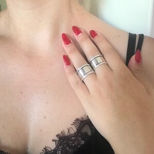 May include: Two silver rings with a wide band design. The rings are worn on the middle and ring fingers of a hand with red nail polish.
