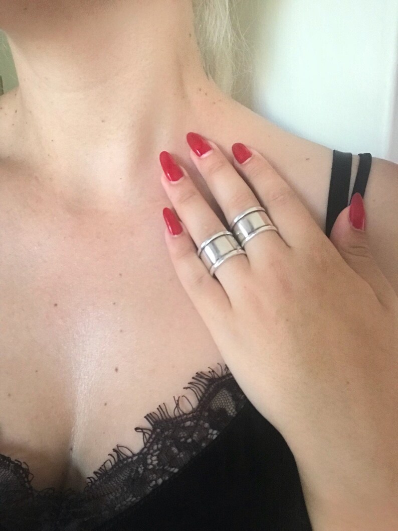 May include: Two silver rings with a wide band design. The rings are worn on the middle and ring fingers of a hand with red nail polish.