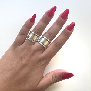 May include: Two silver rings with a wide band design. The rings are worn on the index and middle fingers of a hand with red nail polish.