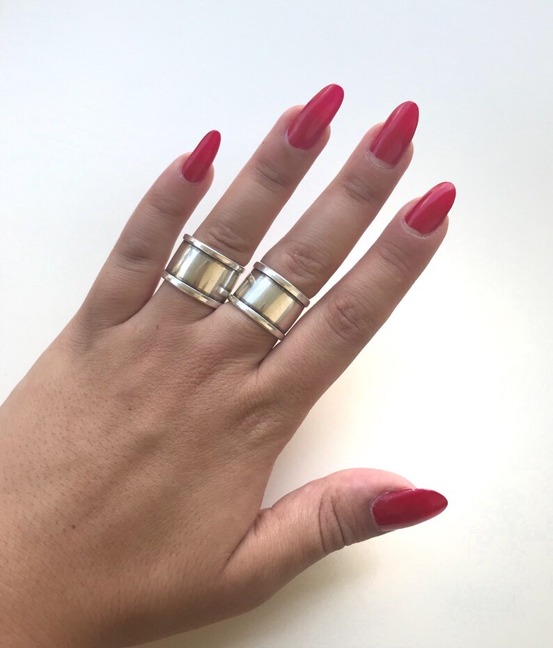 May include: Two silver rings with a wide band design. The rings are worn on the index and middle fingers of a hand with red nail polish.