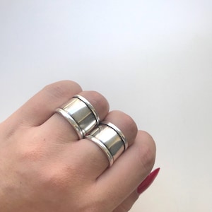 May include: Two silver rings with a wide band and a slightly raised edge. The rings are worn on the index and middle fingers of a hand.