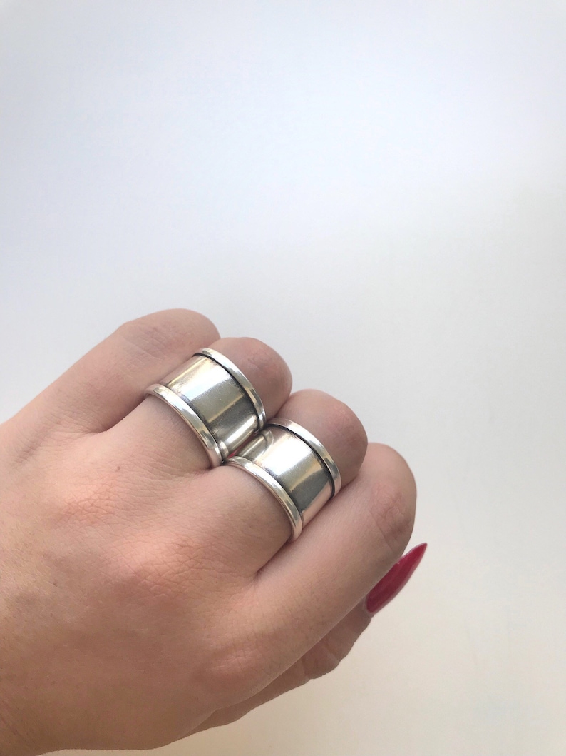 May include: Two silver rings with a wide band and a slightly raised edge. The rings are worn on the index and middle fingers of a hand.