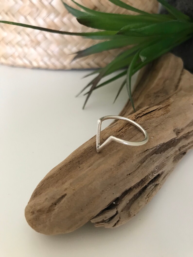 K&ouml;nnte beinhalten: Ein silberner Ring mit einem spitzen, V-f&ouml;rmigen Design. Der Ring liegt auf einem St&uuml;ck Treibholz.