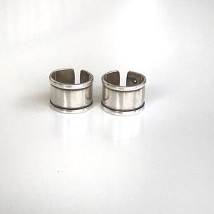 May include: Two silver rings with a wide band and a thin line around the top and bottom of the band.
