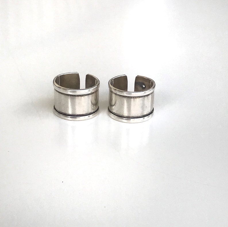 May include: Two silver rings with a wide band and a thin line around the top and bottom of the band.