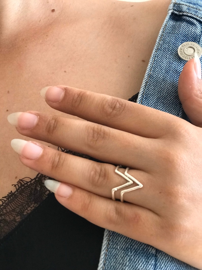 K&ouml;nnte beinhalten: Ein silberner Ring mit einem doppelten Chevron-Design. Der Ring wird am Zeigefinger einer Hand getragen.